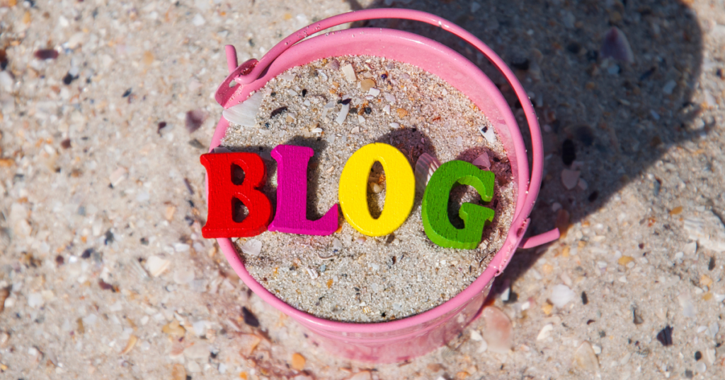 Pink bucket on a sandy beach with felt letters spelling out the word "BLOG" in red, pink, yellow, and green. 