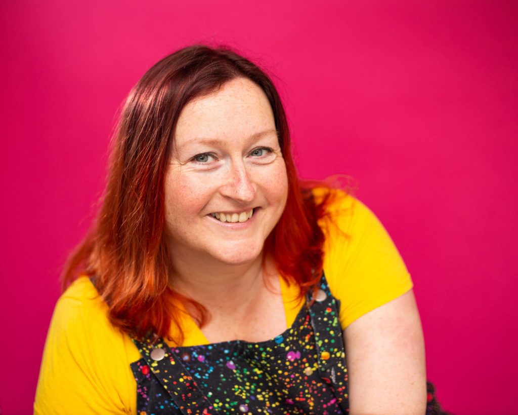 Erin Buck from BizzyBee in a yellow top and dungarees, smiling at the camera with a pink background.
