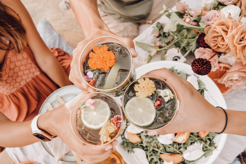 3 women holding a rose gold glass, full of flowery cocktails. Peach, pink, and white flowers on the table.