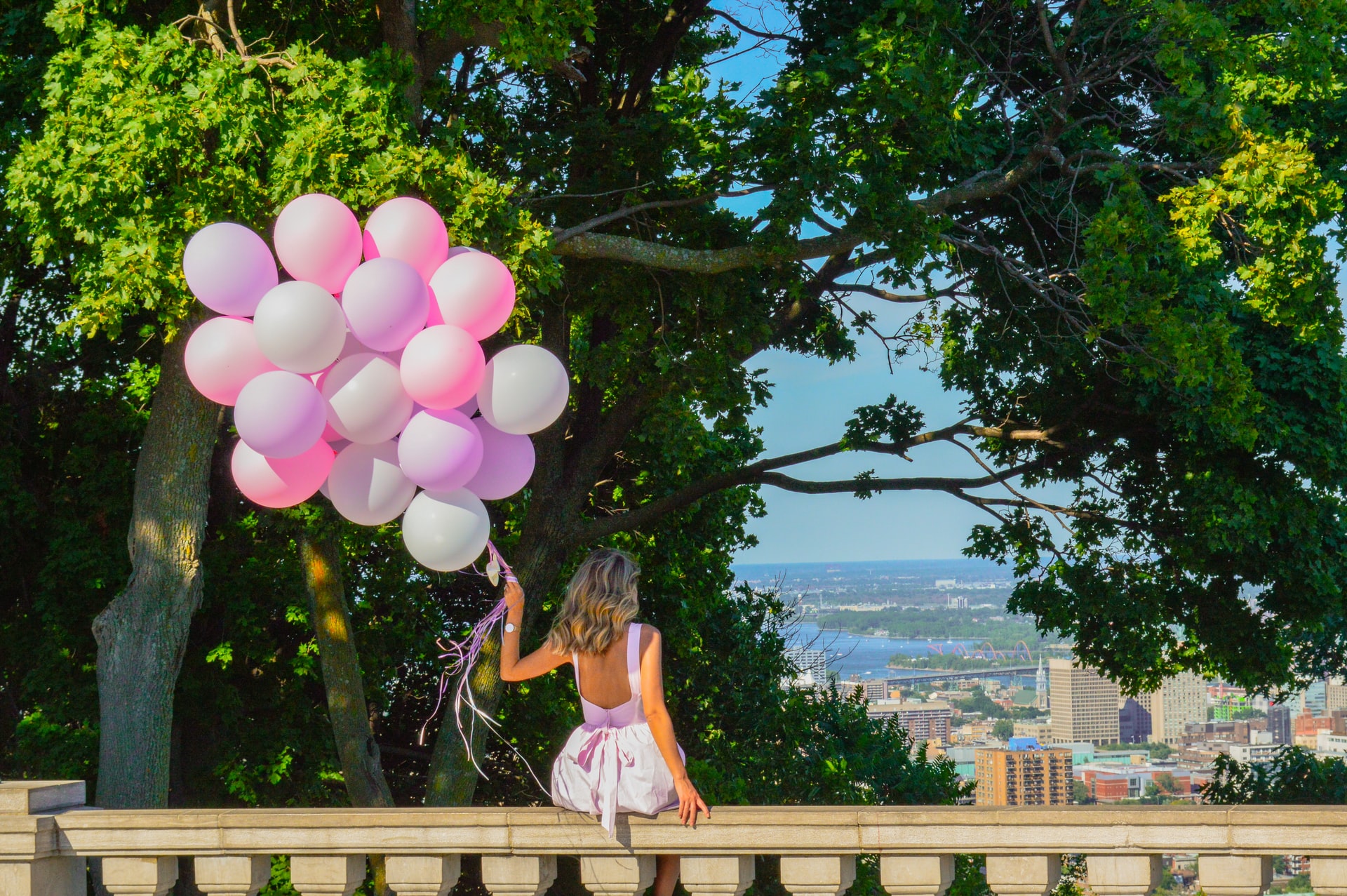 Woman in a dress holding a bunch of pink balloons, overlooking the city.