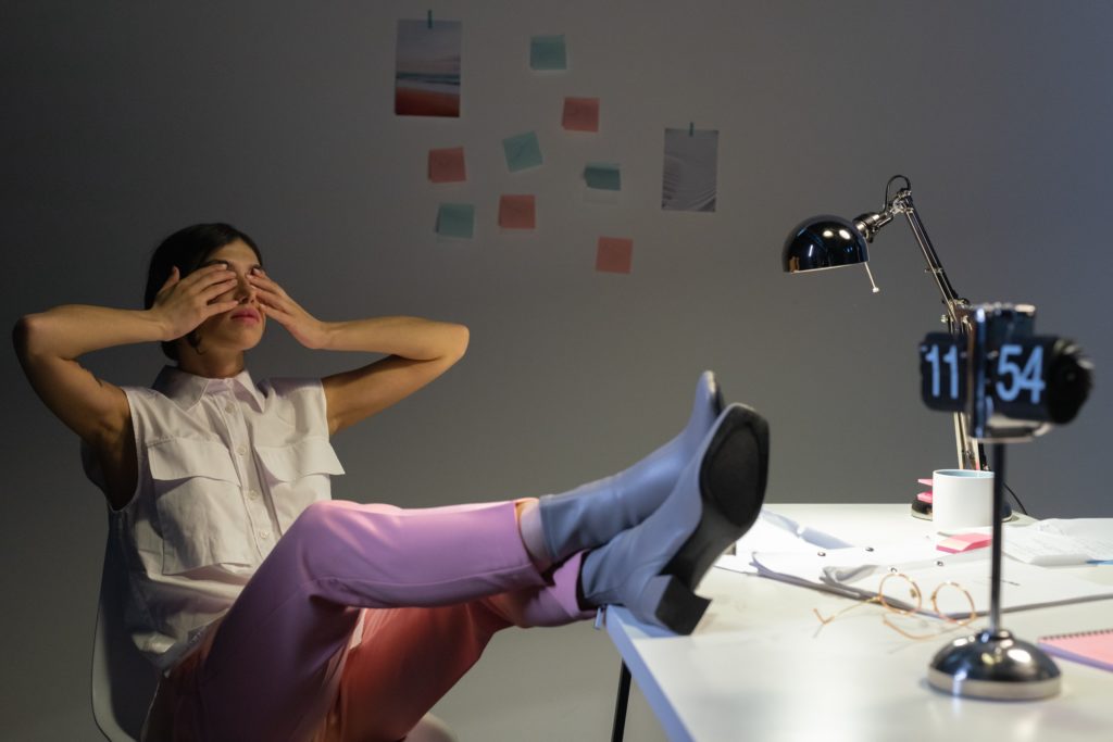 Woman in purple trousers and blue boots exasperated, covering her face with her hands after being exhausted and overworked.