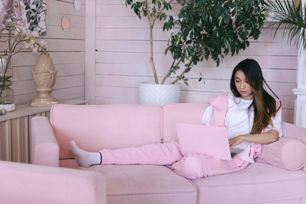Young woman on pink sofa typing on her pink laptop