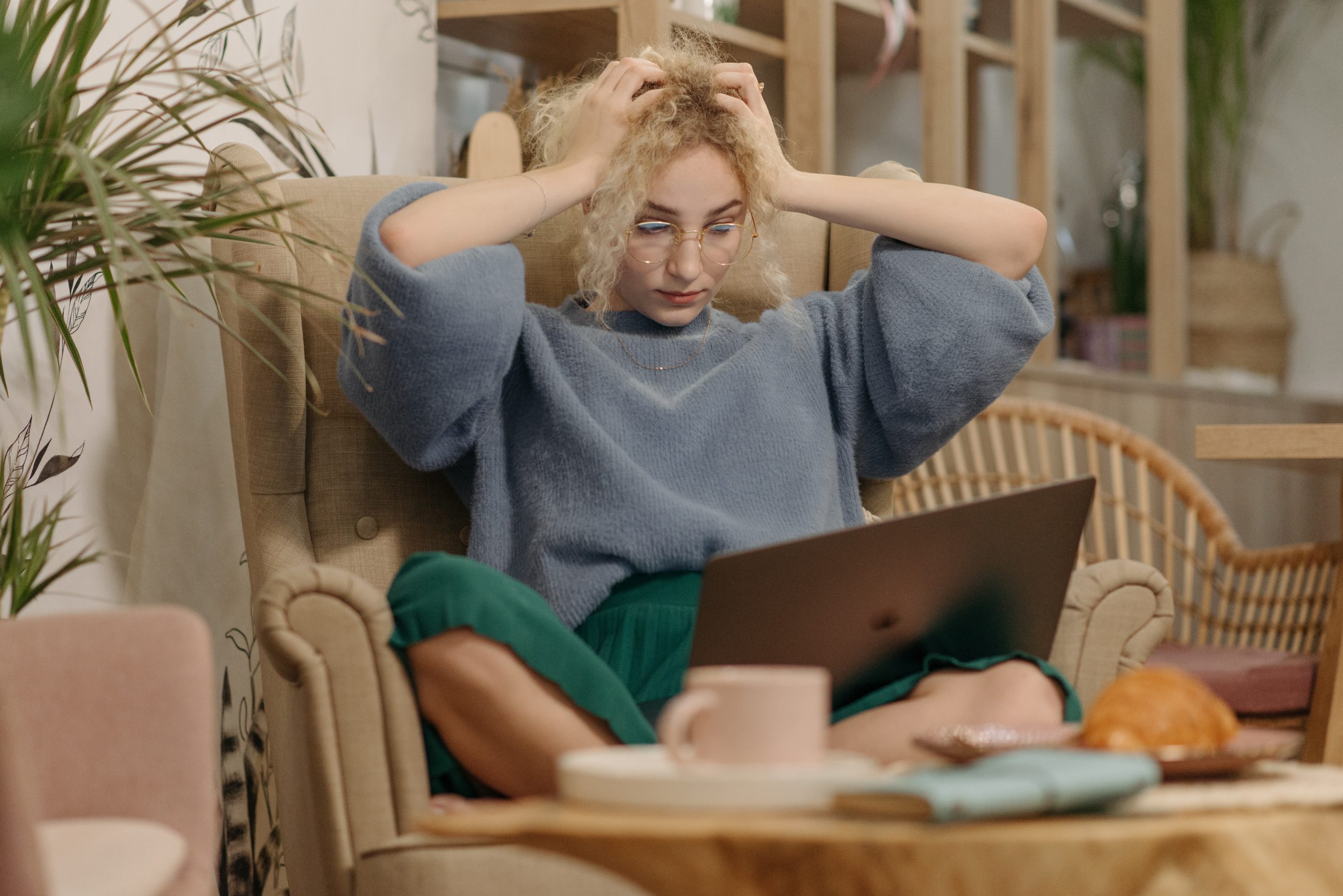 Blonde woman with hands on her head in exasperation, looking at her laptop on a wicker sofa. Distressed.