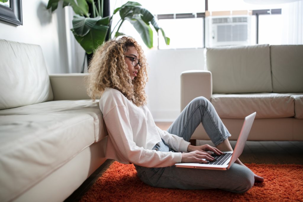 Woman with blonde, curly hair sitting on the floor next to her sofa, working on her laptop.