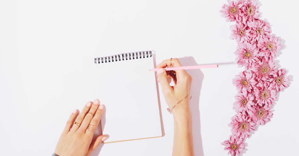 Woman's hands holding a pink pencil against an open notebook next to a collection of pink flowers. 