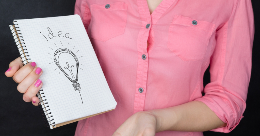 Woman in a salmon pink top with pink nails holding an open sketchbook with a drawing of a lightbulb. 