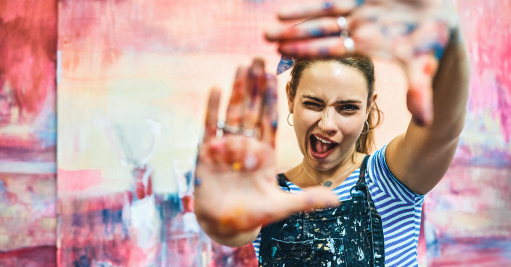 Woman in dungarees splattered with paint using her hands to create a frame at the camera. 