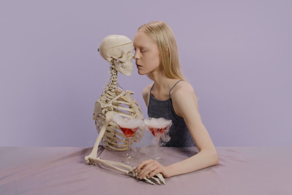 Blonde woman on a date and holding hands with a skeleton having red cocktails with smoke.