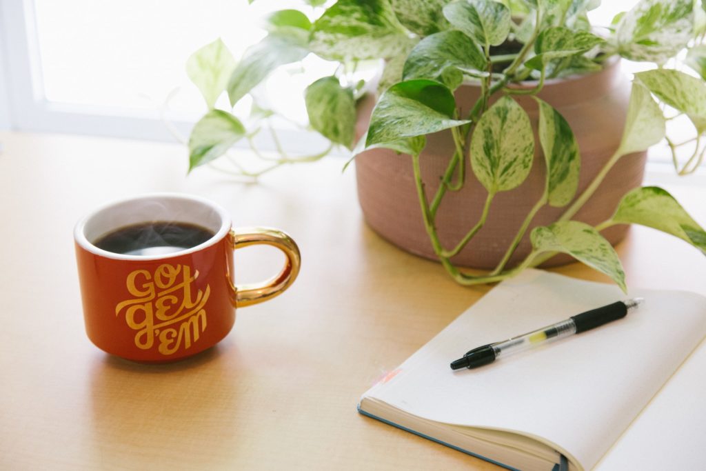 Open notebook and black biro pen, a plant pot with green foliage, a red mug with gold font that says "go get 'em" full of hot, steaming coffee. 