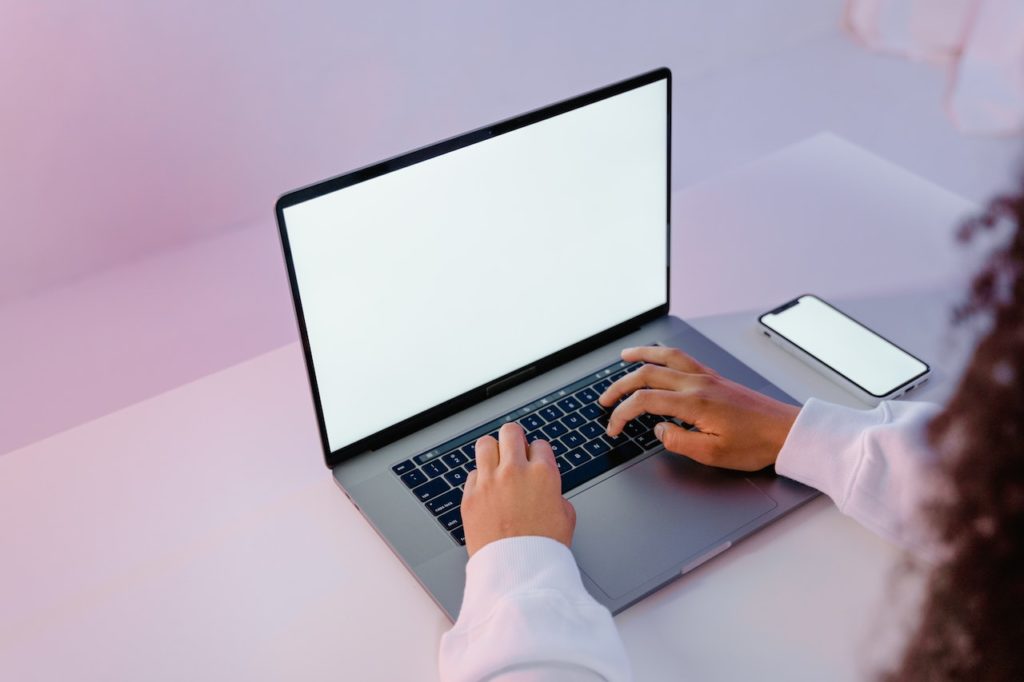 Woman typing on laptop with purple pink background, phone next to her.
